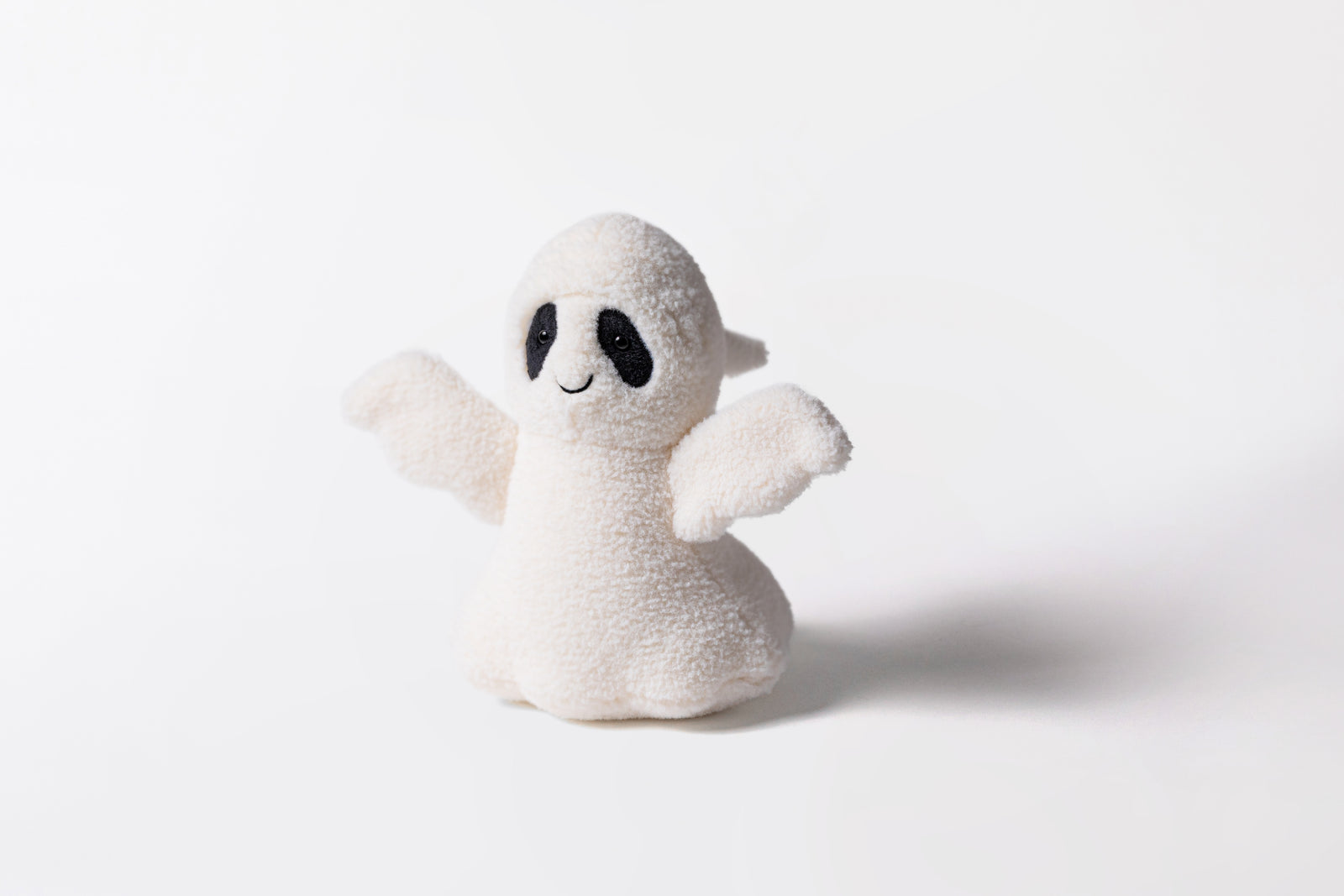 A white plush toy with black patches around it's eyes in the shape and style of a ghost. The ghost wears a smile on it's face. 