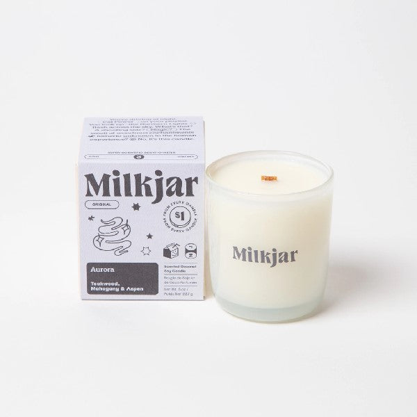 Canadian company Milkjar candle and its box packaging. The white candle is in a translucid glass with "Milkjar" written in dusty purple. The other side of the candle has a cute mist-and-stars doodle in dusty purple. The packaging is a small lilac box with dusty purple text and graphics. 