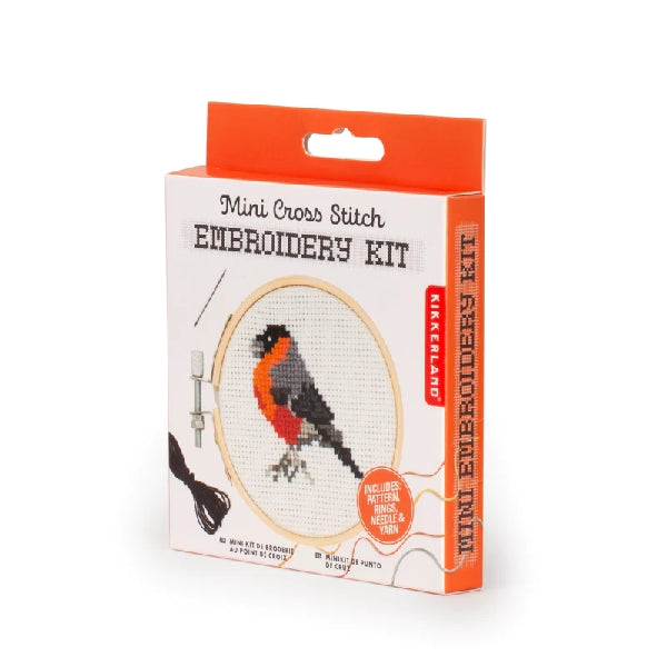 white and orange product box with a hangable tab. the cover of the package shows an embroidered robin at the centre of a wooden circle. main text reads "mini corss stitch embroidery kit". kikkerland product on whit background
