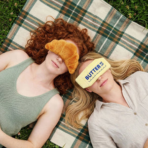 Image of two women wearing food themed eye masks. One wears a yellow one in the shape of a stick of butter with the text 'Butter me up' printed on it, while the other wears a croissant shaped one. Both women are lying on a picnic blanket on the grass. 