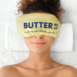 Image of a woman wearing a yellow comfort eye mask in the shape of a stick of butter on a white background. The text 'Butter Me Up' is shown on the eye mask. 
