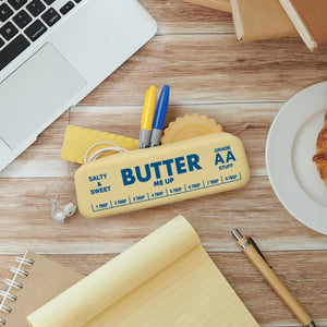 Yellow pouch in the shape and style of a stick of butter. The text 'Butter me up' is printed on it, along with 'Salty & Sweet', 'Grade AA stuff' and some measurements. The pouch is shown lying on a wooden table, along with a plate, a notebad, pens, sticky notes and a laptop.