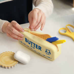 Person opening a yellow pouch in the shape and style of a stick of butter. The text 'Butter me up' is printed on it, along with 'Salty & Sweet', 'Grade AA stuff' and some measurements. The pouch is shown on a white surface beside other objects. 