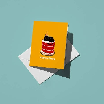 A greeting card featuring an illustration of a black cat sitting on top of a three-tiered red and white birthday cake on a yellow background. A candle sits on the cat's head. The card lies on a white envelope which sits on a blue background. 