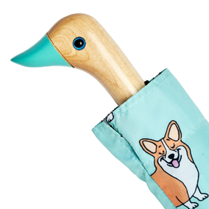 Close up view of the top of a blue umbrella with a corgi print and a wooden duck head shaped handle on a white background.