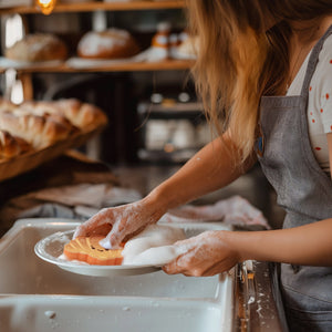 Image of a person using a smiling croissant-shaped kitchen sponge to wash a dish over a sink.