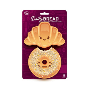 Two smiling bread-themed kitchen sponges in their packaging. One is in the style of a croissant, while the other is a bagel. The purple packaging shows the text 'Daily Bread', the company branding and shows some illustrations of kitchen appliances in the background.
