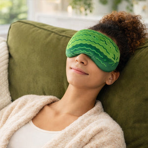 Image of a woman lying on a green couch, wearing a green plush pickle-shaped comfort eye mask over her eyes.
