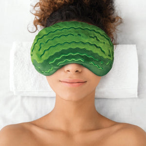 Image of a woman wearing a green plush pickle-shaped comfort eye mask over her eyes on a white background. 