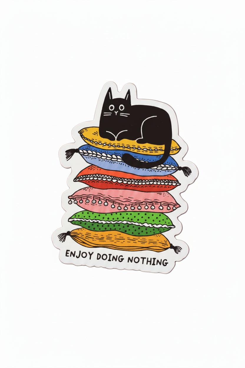 A vinyl sticker featuring a black cat lounging on top of a colorful stack of pillows, with the text 'Enjoy doing nothing' printed below, shown on a solid white background.