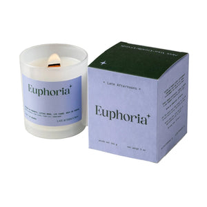 A candle in a milky-white glass jar filled with white wax and a lit wick. The candle sits next to a purple and green packaging box on a white background. The text 'Euphoria' is printed on both the candle and box, along with the company branding. 