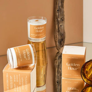 Two candles in milky-white glass jars filled with white wax. One candle sits on top of an orange and cream packaging box while another is placed on top of an upturned orange glass. Another packaging box sits across the surface from the others. The text 'Golden Hour' is printed on the candles and boxes, along with the company branding.  
