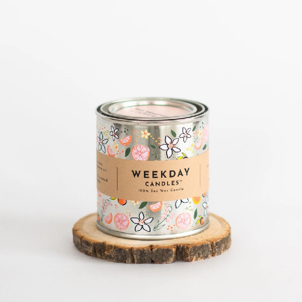 silver tin candle with painted doodles of flowers and citrus fruits. a brown tag wraps the tin. on it, "Weekday Candles 100% Soy Wax Candle", the name of the product, the ingredients and instructions are written in a sharp black font.