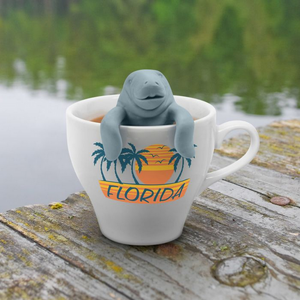 Image of a manatee-shaped tea infuser hanging inside a white mug filled with tea. The mug sits on a wooden surface.