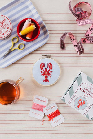 Cross-stitched lobster design contained in a wooden embroidery hoop sits on top of a striped surface along with it's packaging, a cup of tea and crafting materials.