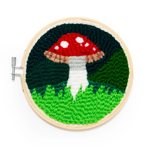 bright punch needle mini tapestry on a circular wooden frame. a red-cap mushroom on grass and a dark green background. kikkerland product on white background