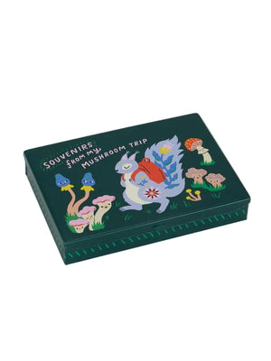 Image of a dark green pocket-sized storage tin featuring an illustration of a a squirrel wearing a red backpack walking on grass, beside smiling mushrooms. The text 'Souvenirs from my mushroom trip' is printed over the images. The tin sits on a white background. 