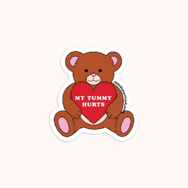 white die-cut sticker of a smiling teddy bear holding a red heart with the text "my tummy hurts"