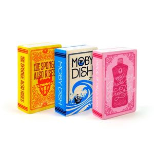 Image of a three-pack of kitchen sponges with book themes. One is pink with imagery and the text 'Pride & Prejudish', another is yellow with orange imagery and the text 'The Sponge Also Rises', and the other is white with blue imagery and the text 'Moby Dish'. 