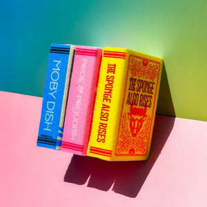 Side view of three kitchen sponges with book themes. The one at the front is yellow with orange illustrations and the text 'The Sponge Also Rises' is shown both on the front and along the spine. The one behind it is pink with 'Pride & Prejudish' along the spine, and behind it a blue spine with "Moby Dish' written on it.