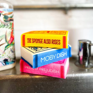 Image of a stack of three kitchen sponges with book themes sitting on a sink. One has 'The Sponge Also Rises' written along the spine, another has 'Moby Dish', while the other has 'Pride & Prejudish'.