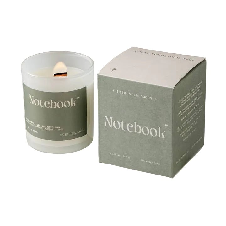 A candle in a milky-white glass jar filled with white wax and a lit wick. The candle sits next to a dark green and light green packaging box on a white background. The text 'Notebook' is printed on both the candle and box, along with the company branding. 