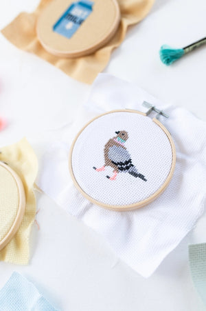 Cross-stitched pigeon design in an embroidery hoop on a white surface.