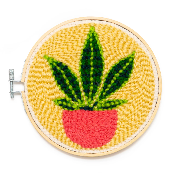 bright punch needle mini tapestry on a circular wooden frame. a plant in a pink planter is on a yellow background. kikkerland product on white background