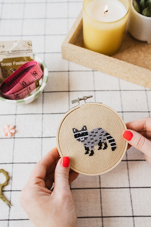 Person holding a cross-stitched racoon design contained within a circular embroidery hoop on a checkered tablecloth.