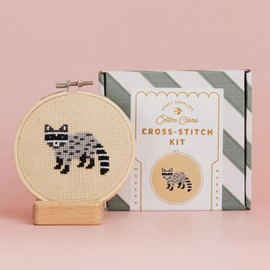 A cross-stitch kit with a raccoon design contained within a circular embroidery hoop stands beside it's packaging on a pink background.