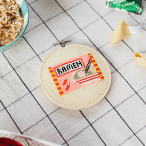 Cross-stitched ramen themed design on a small hoop with a bowl of ramen in the background.