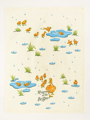 Open tea towel featuring llustrations of a duck family walking in the rain and playing in puddles, on a white background. The text 'Ringmaster of the Shit Show!' is printed in the foreground. 