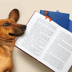Image of a brown and black dachshund-shaped bookmark perched on top of a book page. The open book lies on a flat surface beside a real dachsund dog. 