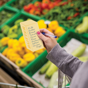 Image of a person holding sticky notes that look like a yellow bar of butter. The person also holds a pen and a grocery list is written on the sticky note. The text 'Big 'ol butter' is printed along the side of the sticky notes. Fruit and vegetables are shown in the background.