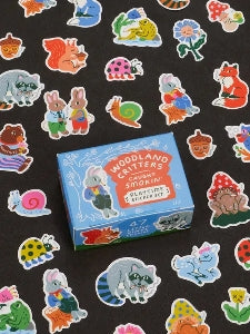 Image showing a packaging box for a Woodland Critters Caught Smoking themed sticker set. Stickers of colourful woodland creatures surround the box. The box and stickers sit against a black surface.