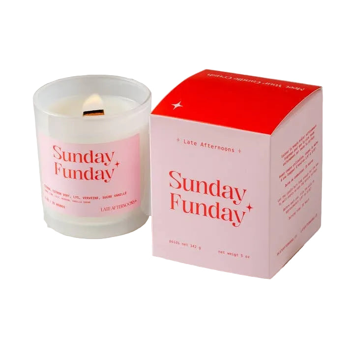 A candle in a milky-white glass jar filled with white wax and a lit wick. The candle sits next to a pink and red packaging box on a white background. The text 'Sunday Funday' is printed on both the candle and box, along with the company branding. 