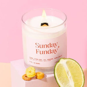 A candle in a milky-white glass jar filled with white wax and a lit wick. The text 'Sunday Funday' is printed on a light pink coloured label on the candle, along with the company branding. The candle sits against a pink background, along with half a lime and some fruit loops.