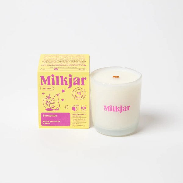 Canadian company Milkjar candle and its box packaging. The white candle is in a white glass with "Milkjar" written in pink. The other side of the candle has a cute kissing pear-and-nectarine-couple doodle in pink. The packaging is a small pale yellow box with pink text and graphics.