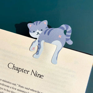 Image of a grey striped cat-themed bookmark perched on the page of a book, against a blue background.