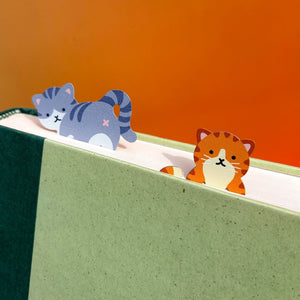 Image of two cat-themed bookmarks perched on top of book pages. One cat is grey striped while the other is orange striped. 