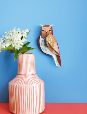 3D owl wall art hanging on a blue background beside a striped vase containing flowers. The owl is brown, white and blue in colour.