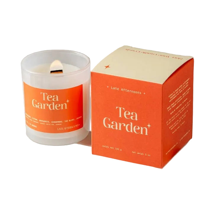 A candle in a milky-white glass jar filled with white wax and a lit wick. The candle sits next to an orange and cream packaging box on a white background. The text 'Tea Garden' is printed on both the candle and box, along with the company branding. 
