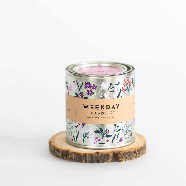 silver tin candle with painted doodles with of white, purple, pink, lilac flowers and dark stems. a brown tag wraps the tin. on it, "Weekday Candles 100% Soy Wax Candle", the name of the product, the ingredients and instructions are written in a sharp black font.
