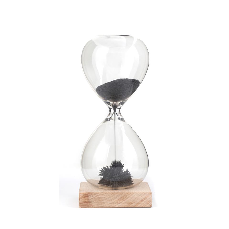 hourglass with black magnetic sand flowing from one bulb to the next. the sand at the bottom forms magnetic spikes. the glass sits on a light wooden block. kikkerland product on white background
